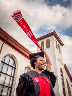 Student in graduation gown waving sash in front of the School of Communication and Media